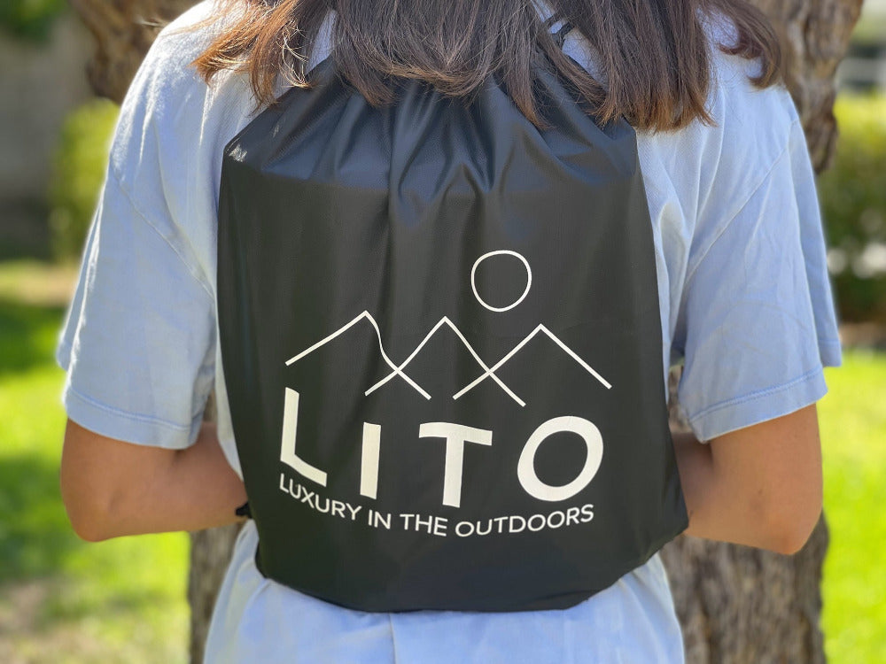LITO black backpack with white lettering saying LITO luxury in the outdoors worn by a woman with brown hair wearing a white tshirt
