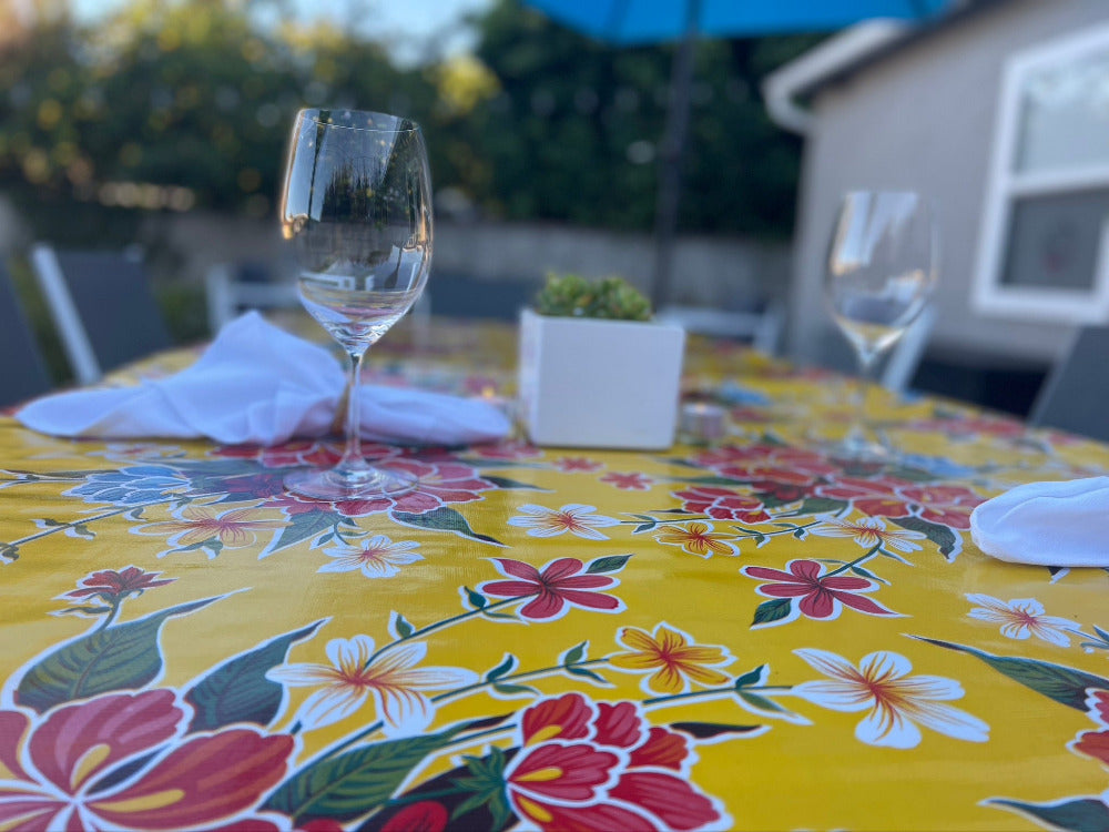 red, yellow and white flower pattern on yellow outdoor tablecloth with planter, two wine glasses and two white napkins