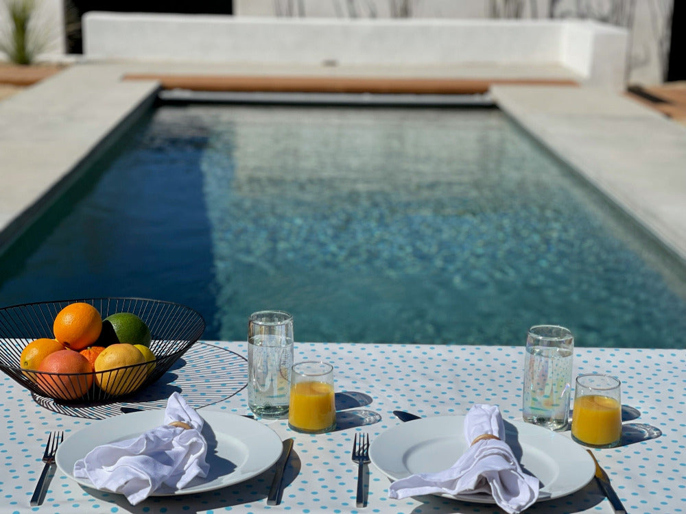 blue and white outdoor tablecloth with fruit bowl, two place settings, two glasses of water and two glasses of orange juice overlooking a pool.
